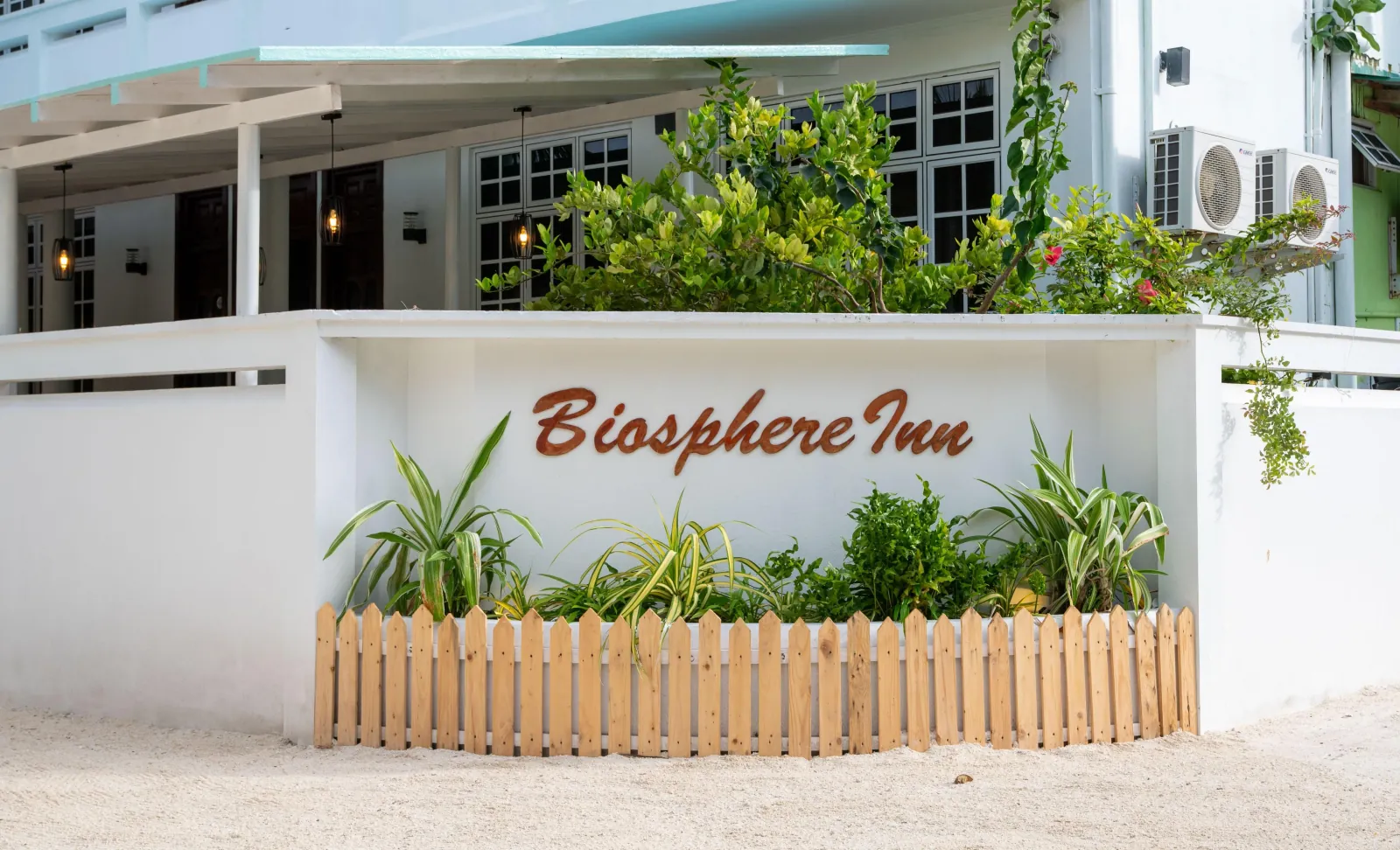 White entrance wall of Biosphere Inn with the guesthouse name in lettering, a picket fence and tropical planters on a sand street