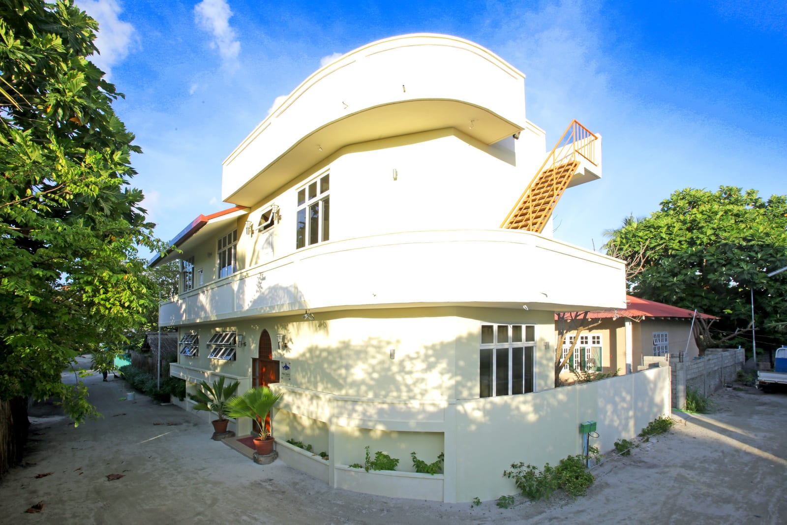 The white two-storey Hanifaru Transit Inn with curved balconies on a sandy lane under blue sky, greenery on either side