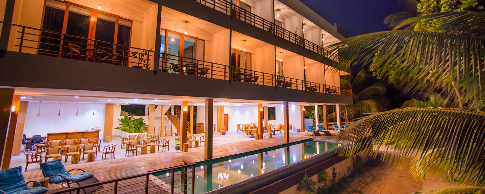 The three-storey Kiha Beach hotel at dusk, balconies lit, with the saltwater lap pool and open-air restaurant deck in the foreground