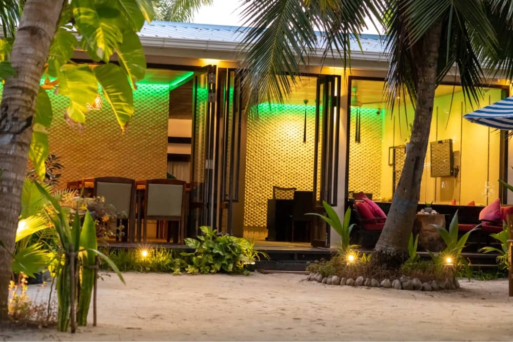 LVIS Blancura's open-fronted beachfront restaurant pavilion lit up at dusk behind palms and white sand